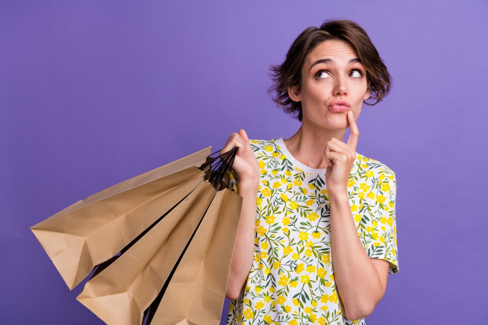 A young woman in a floral shirt holds several empty shopping bags and looks thoughtfully to the side.