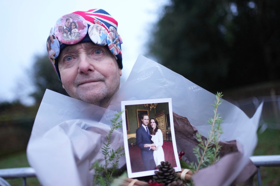 A royal fan wearing a hat adorned with royal family photos holds a photo of Prince William and Kate and a bouquet of flowers while waiting to see the royal family.