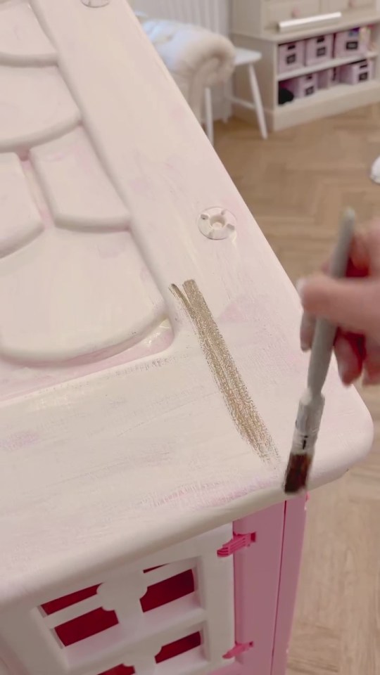 A person painting a pink playhouse white with gold accents.