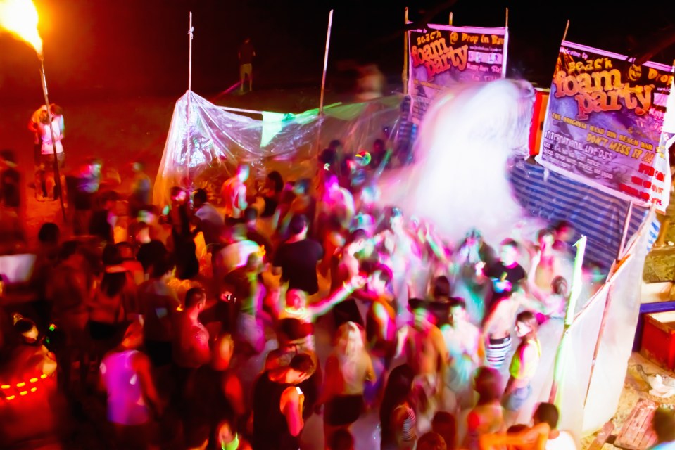 A large group of people dancing at a brightly lit beach foam party at night.