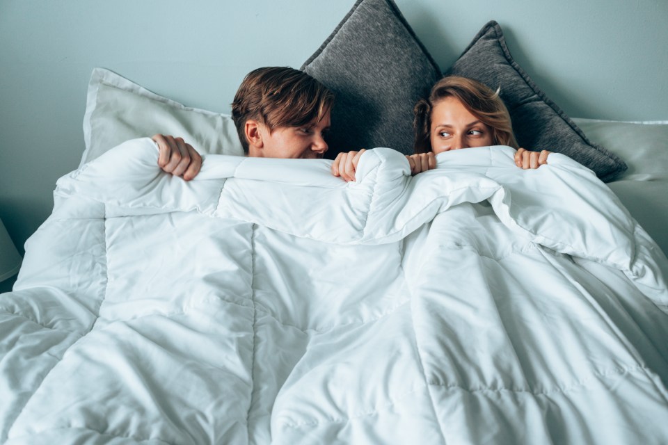 A couple hides under a duvet in bed.