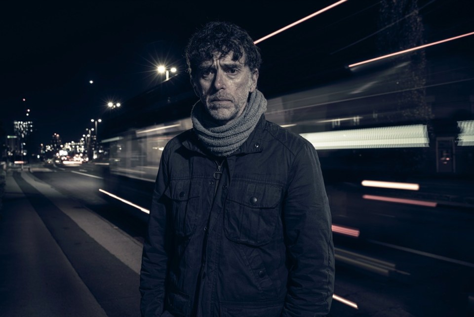 A man stands on a city street at night, looking somber.