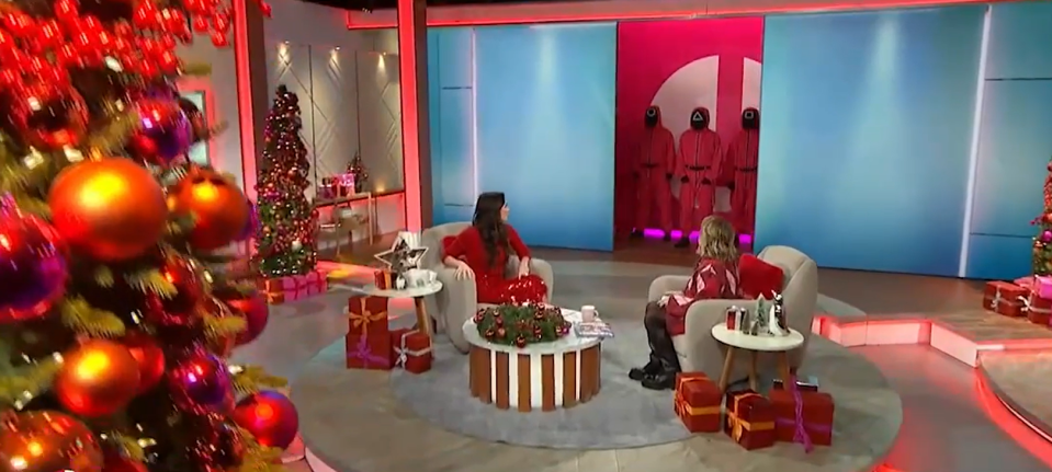 Christine Lampard on a talk show, reacting to an alarm and three Squid Game-costumed figures.