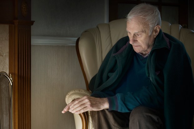 Elderly man sits in a chair, appearing worried near a fireplace.