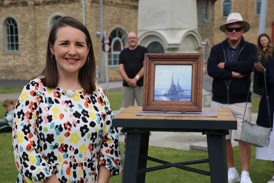 A woman stands smiling in front of a framed painting of sailing ships, displayed on a small table.