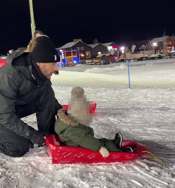 A man helps a child on a red sled in the snow at night.