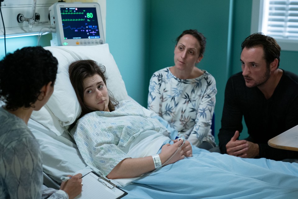 A doctor takes notes while talking to a young woman in a hospital bed, with her parents nearby.