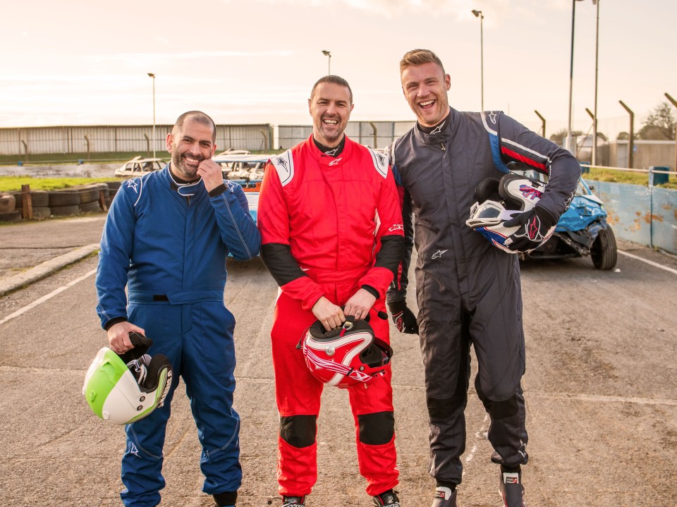 Three men in racing suits stand in a parking lot with helmets, smiling.  A damaged car is visible in the background.