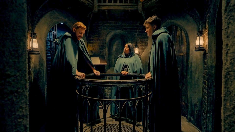 Three people in dark green cloaks stand around a circular structure in a dimly lit room.