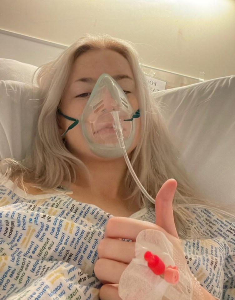 Woman in hospital bed giving thumbs up while wearing an oxygen mask.