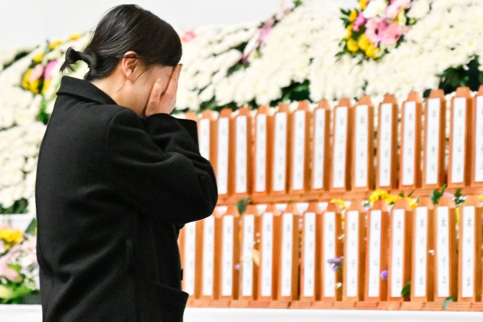 A grieving woman stands before a memorial altar with numerous name plaques.