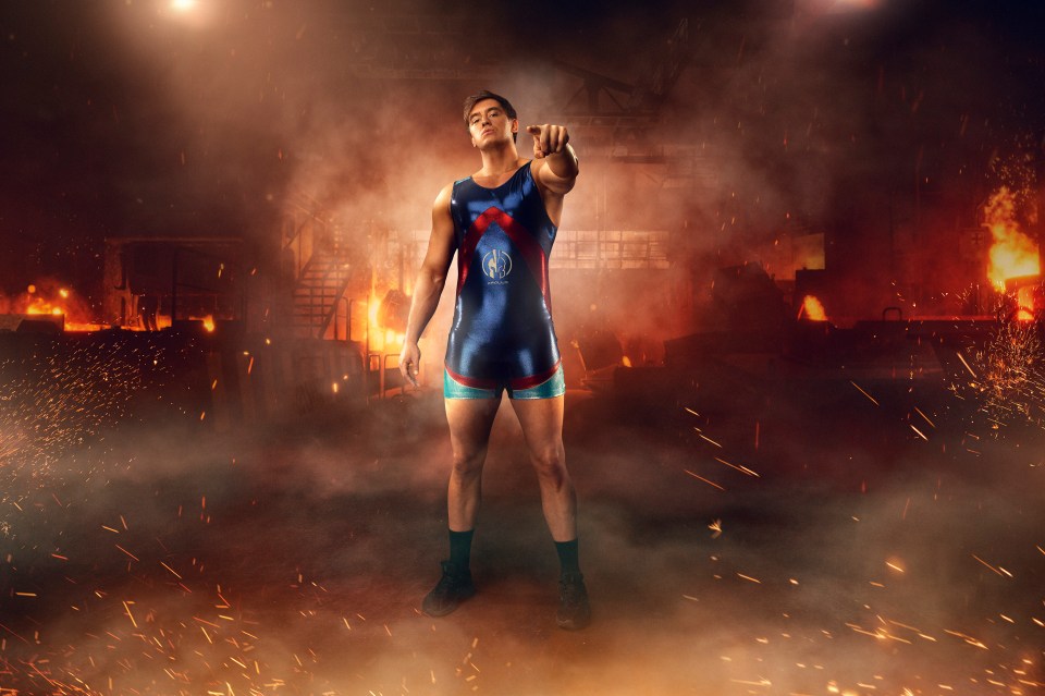 A wrestler in a blue and red uniform points towards the viewer in a fiery industrial setting.