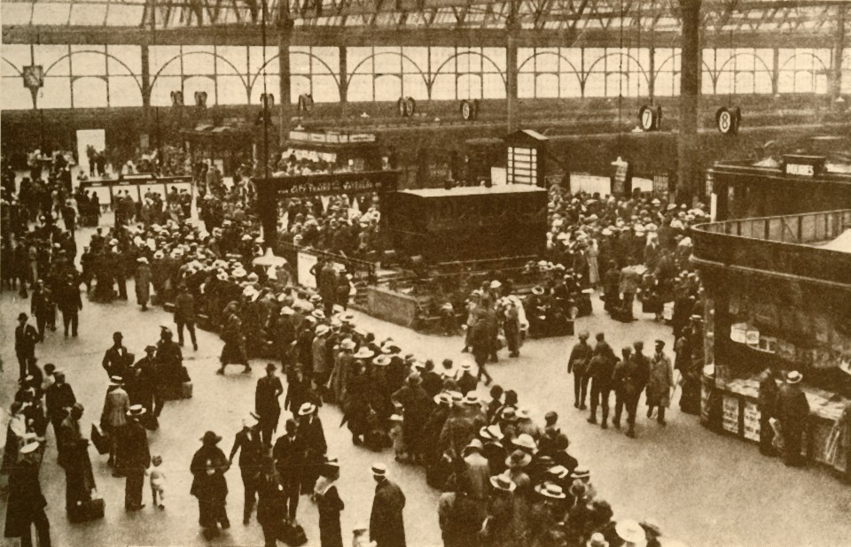 A 1930s black and white photo of a crowded train station concourse, showing a long queue of passengers waiting for trains at Waterloo Station in London.