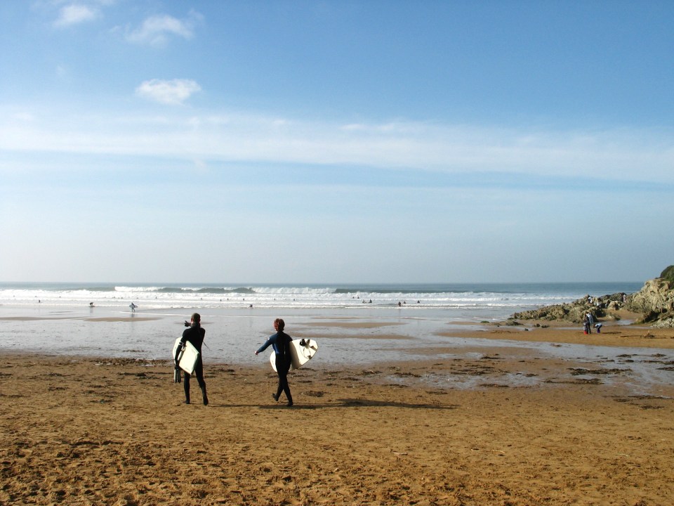 Two surfers carrying surfboards walk on a sandy beach toward the ocean.