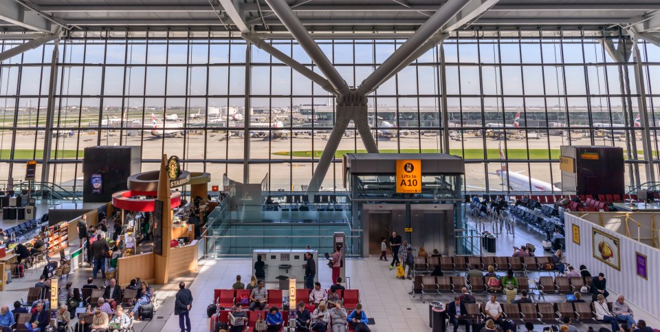 Interior view of Heathrow Terminal 5, showing passengers waiting in the terminal with airplanes visible through large windows.