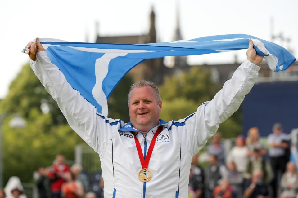 A Scottish lawn bowler holding a gold medal and the Saltire at a medal ceremony.