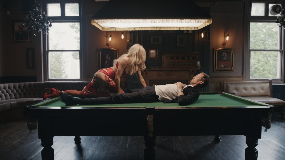 A woman in red lingerie leans over a man lying on a pool table in a pub.