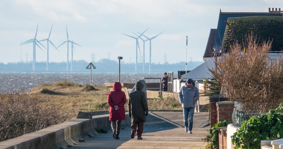 People walking along a seafront path with wind turbines in the distance.