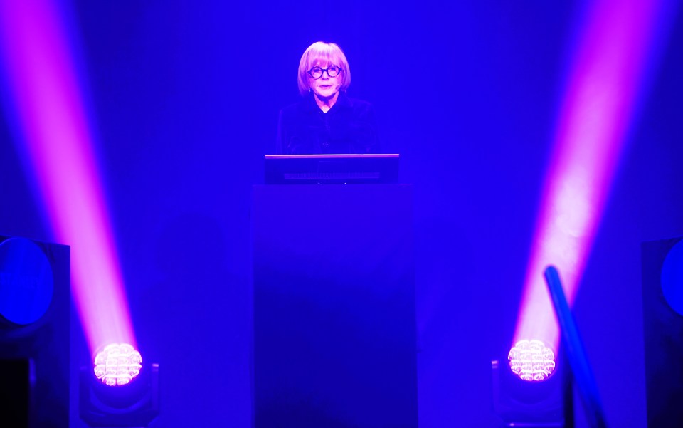 Anne Robinson at a podium under purple stage lights.