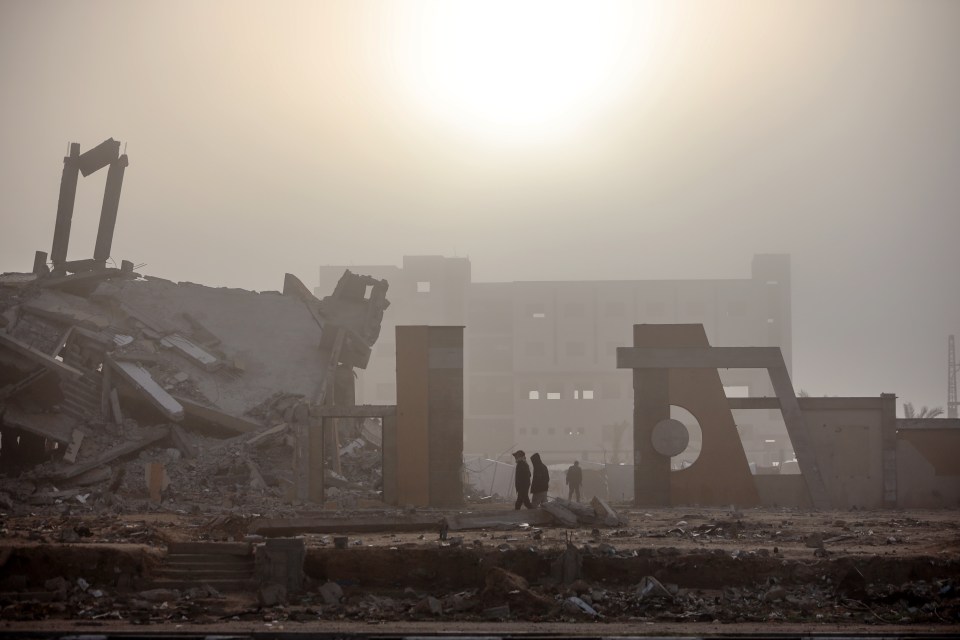 People walk past rubble and damaged buildings in the fog.