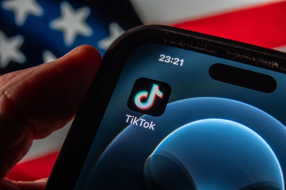 Photo illustration of a smartphone displaying the TikTok app, with a blurred US flag in the background.