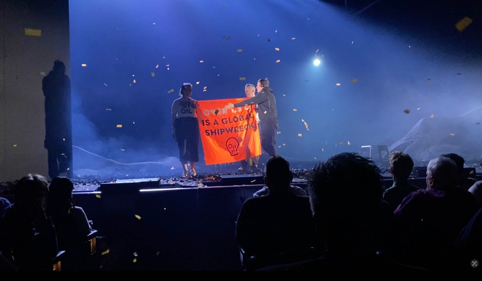 Two protestors holding a banner reading "Over 1.5 degrees is a global shipwreck" on a stage during a theatrical performance.