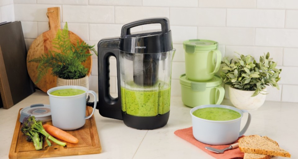 A blender filled with green soup sits on a kitchen counter next to two mugs of soup and food storage containers.