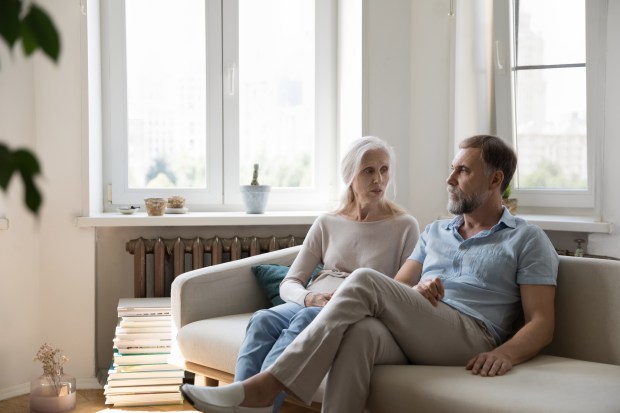 A senior couple sits on a couch, looking concerned.