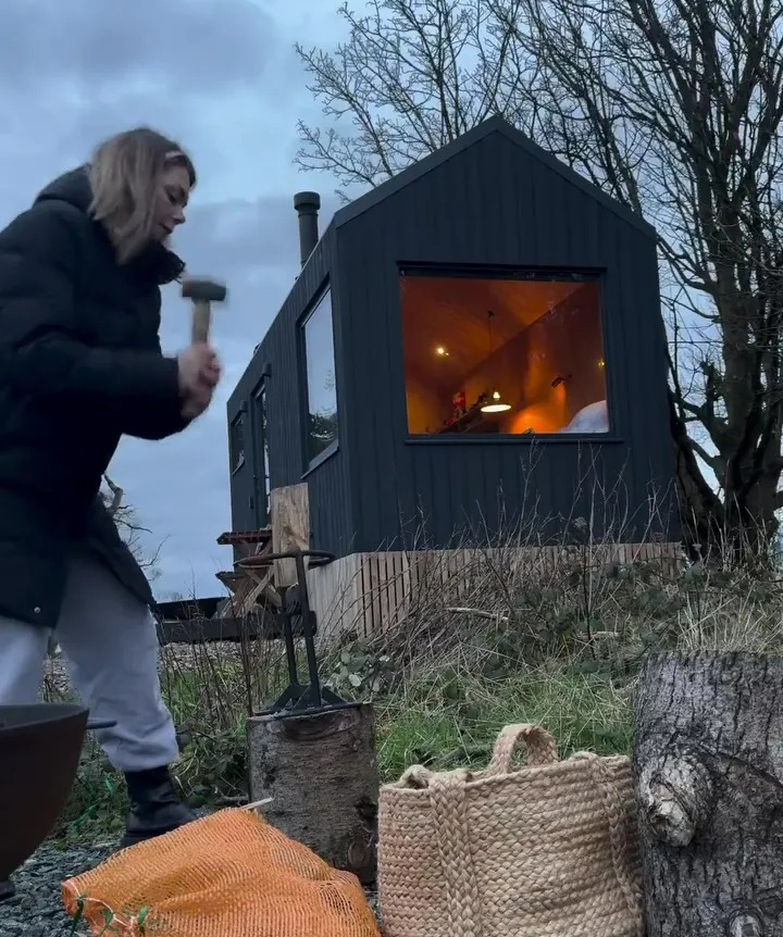 Woman chopping wood outside a dark cabin.