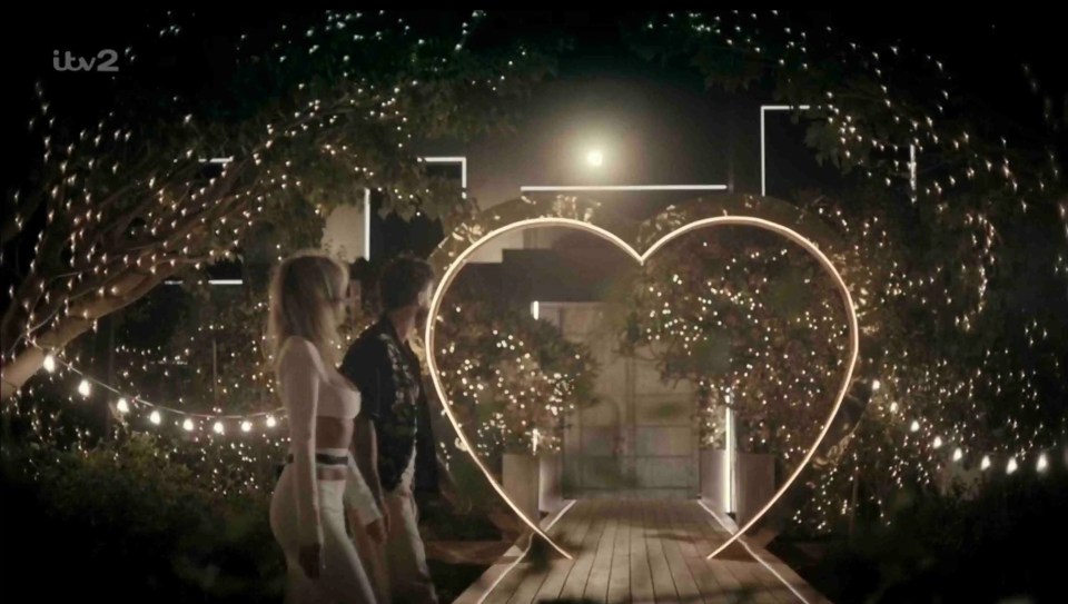 Couple walking through heart-shaped archway at night.