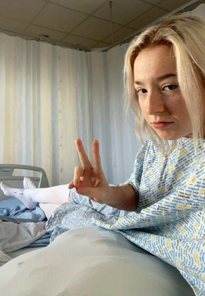 Woman in hospital gown giving peace sign in hospital bed.