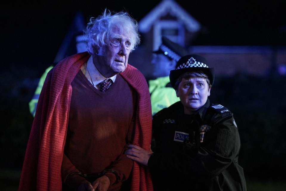 A handcuffed elderly man in a red sweater is escorted by a police officer.