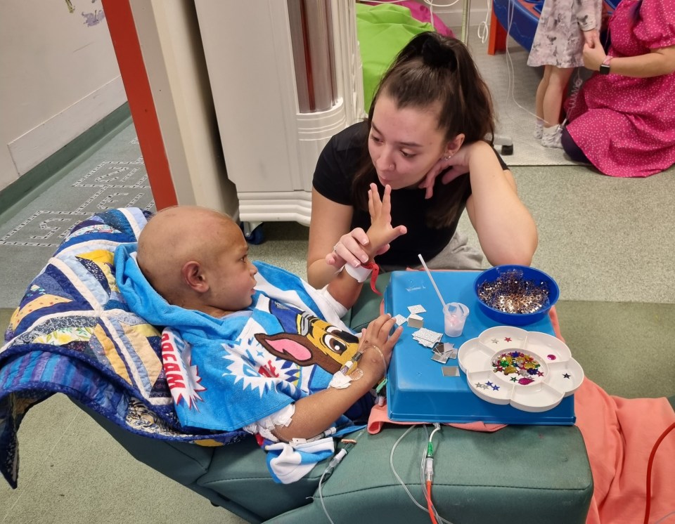 A young girl and her brother, who is undergoing treatment for leukemia, are sitting together in a hospital room.