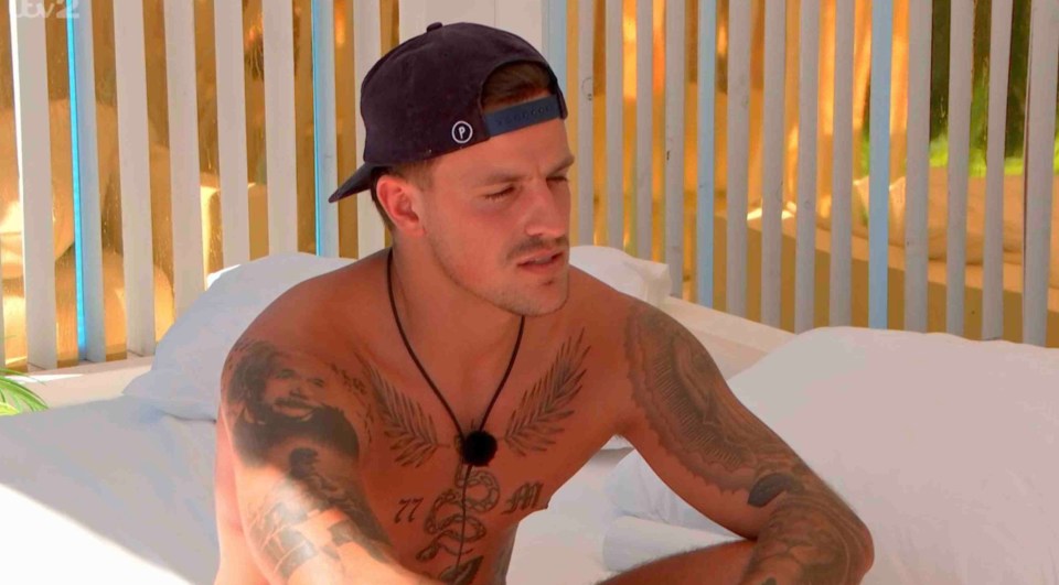 Luca Bish from Love Island sitting on a bed, looking pensive.