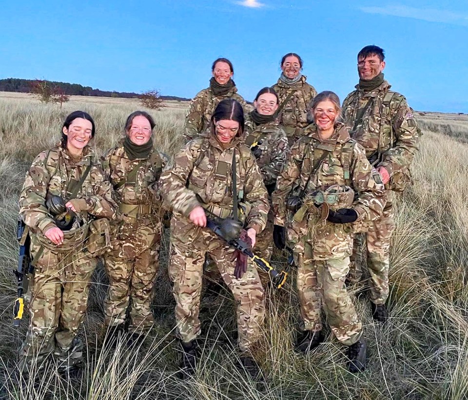 A squadron of soldiers in camouflage uniforms stand in a field at dusk.