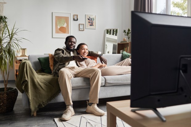 A young Black couple watches TV together on a couch in their living room.