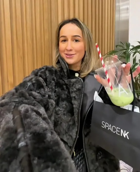 Woman in a fuzzy black jacket holding a green drink and a SpaceNK shopping bag.