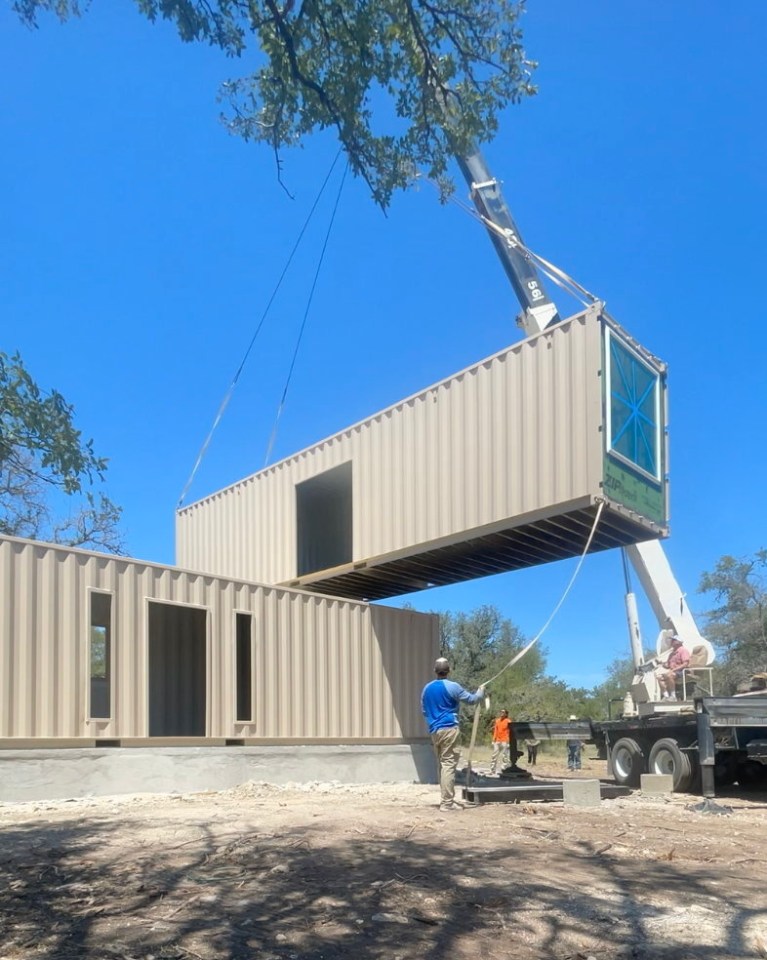 Shipping containers being lifted by a crane onto a foundation during home construction.