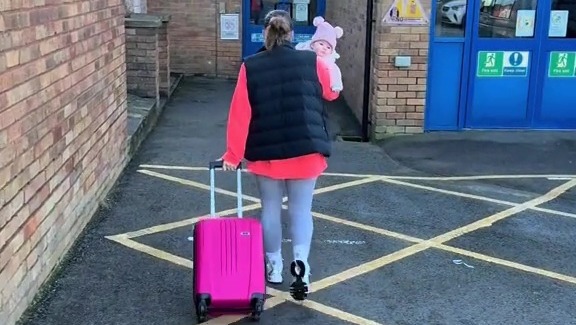 Woman pulling a pink suitcase while carrying a baby, heading towards a swimming pool.