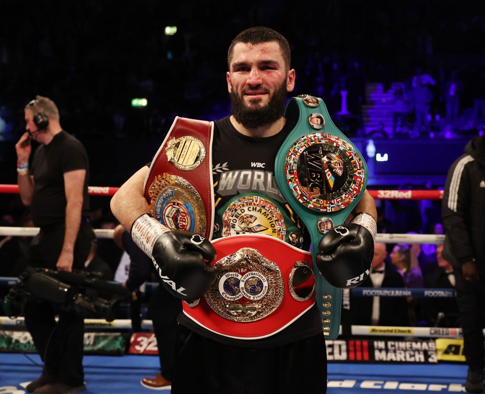 Artur Beterbiev holding WBC, IBF, and WBO light heavyweight championship belts after winning a boxing match.