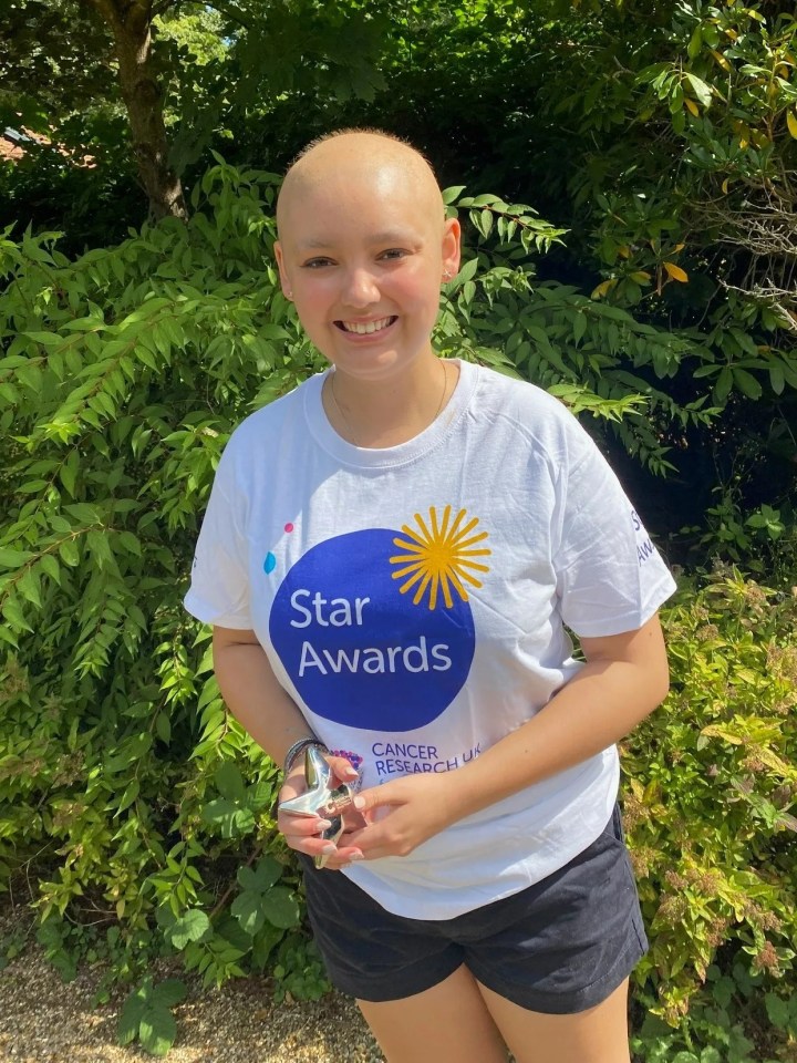 Teenage girl with shaved head holding an award, wearing a Cancer Research UK t-shirt.