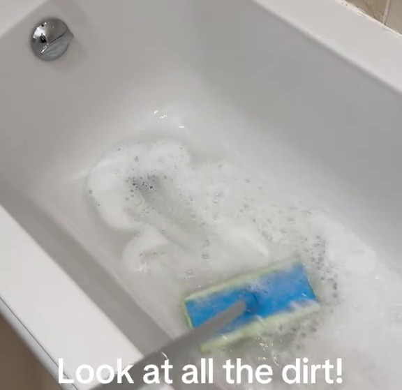 A cleaning tool scrubs a bathtub, revealing significant dirt in the soapy water.