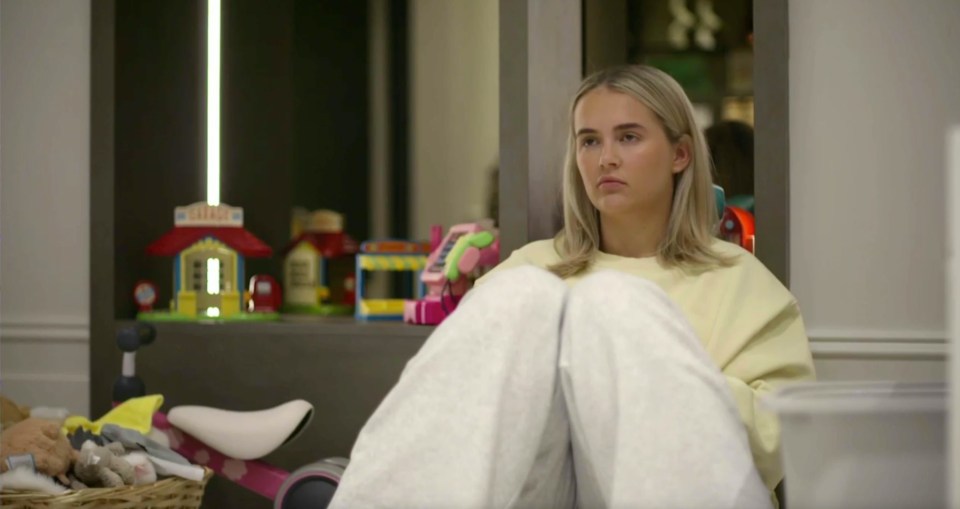 Screenshot of a young woman sitting on the floor, looking pensive; children's toys are visible in the background.