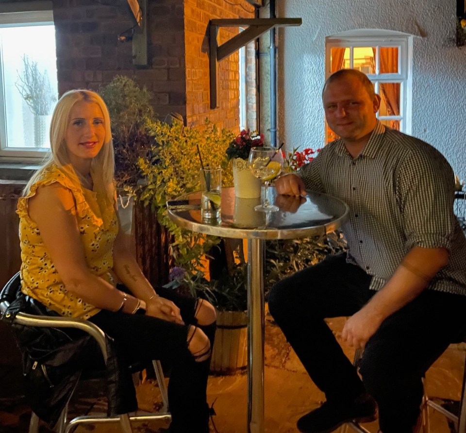 A man and woman sit at a small table outside a pub at night.