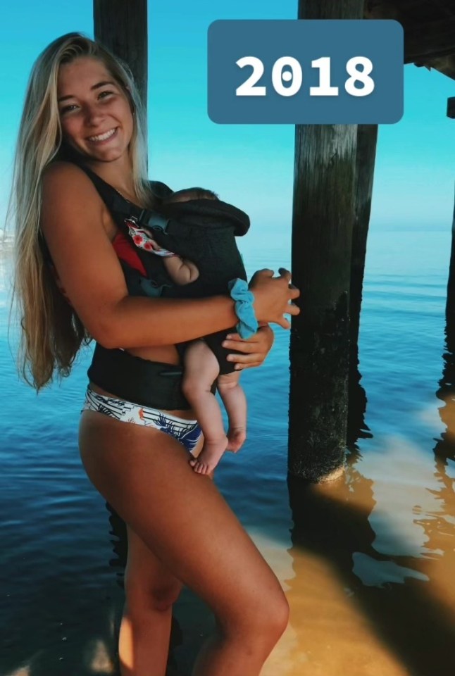 Woman in a swimsuit holding a baby in a carrier at the beach in 2018.