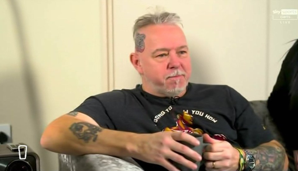 A man with tattoos and graying hair sits and holds a mug.