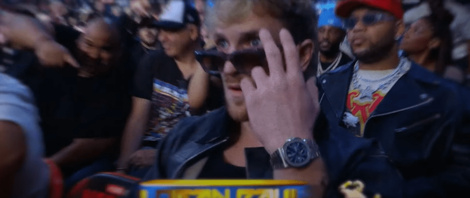 A man in sunglasses adjusts his glasses while sitting in a crowd.