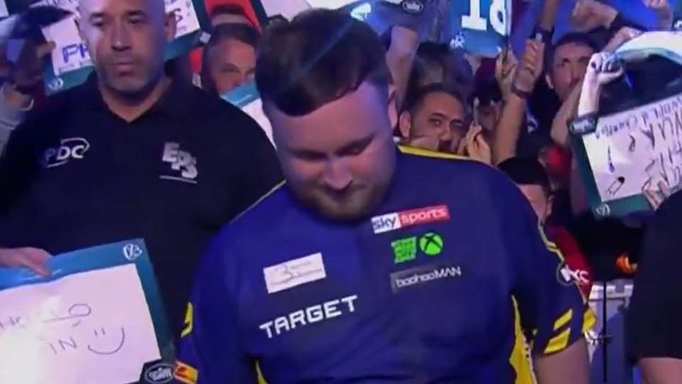 A darts player with his head down, surrounded by spectators holding up signs.