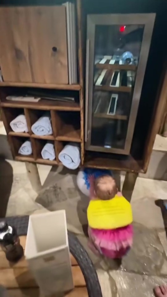 Baby in pink tutu near wooden cabinet with towels and beverage cooler.