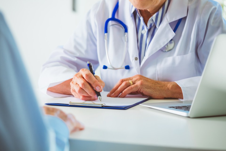 Senior female doctor writing on clipboard during discussion with male patient. Doctor and man sitting at the desk. Close up of hands, unrecognizable people.